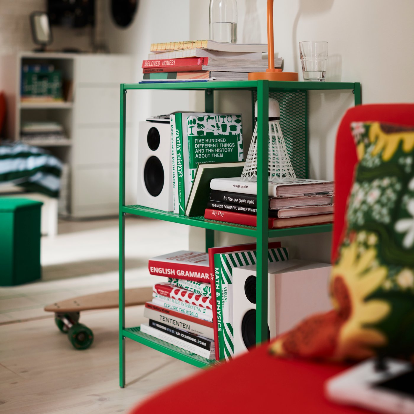 A bright green, metal BAGGEBO shelving unit, filled with books and speakers, next to a skateboard on a wooden bedroom floor.