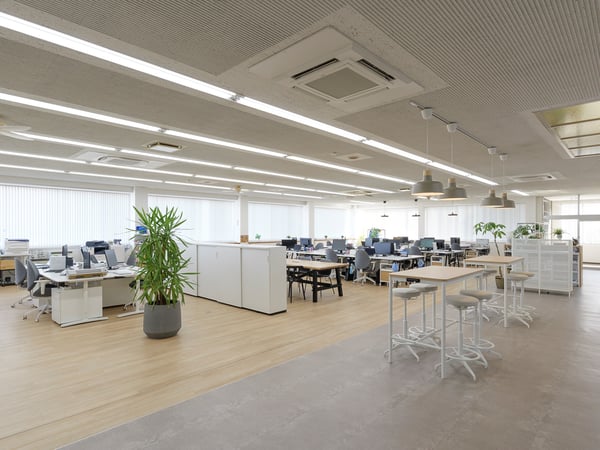 天井まで続く照明と木目フロアが広がるオフィス。デスクエリアとハイテーブル、観葉植物が並ぶ開放的なレイアウト。