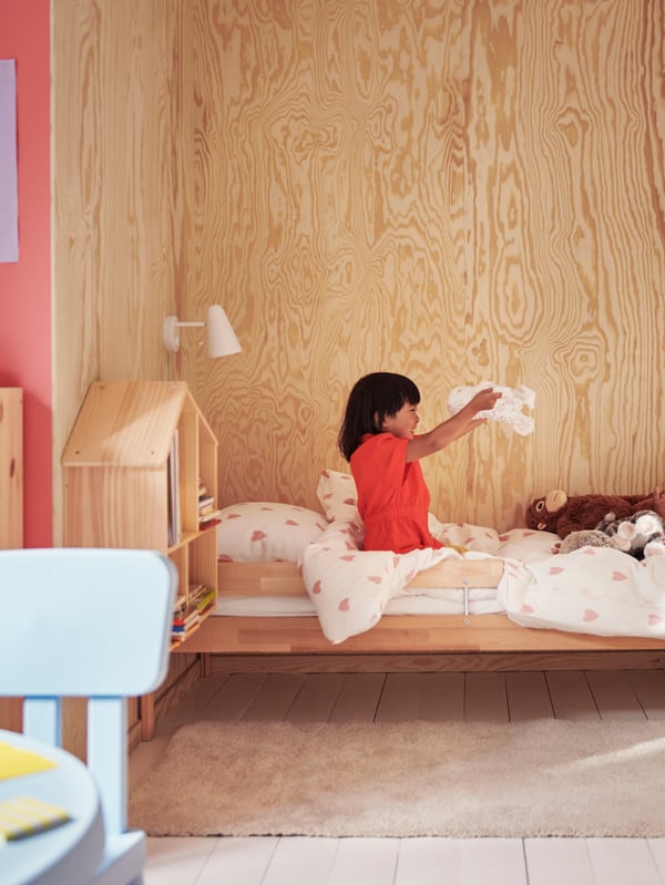 A child sitting on a SNIGLAR bed with guard rail holds a soft toy. White/pink heart BARNDRÖM bed linen covers the bed.