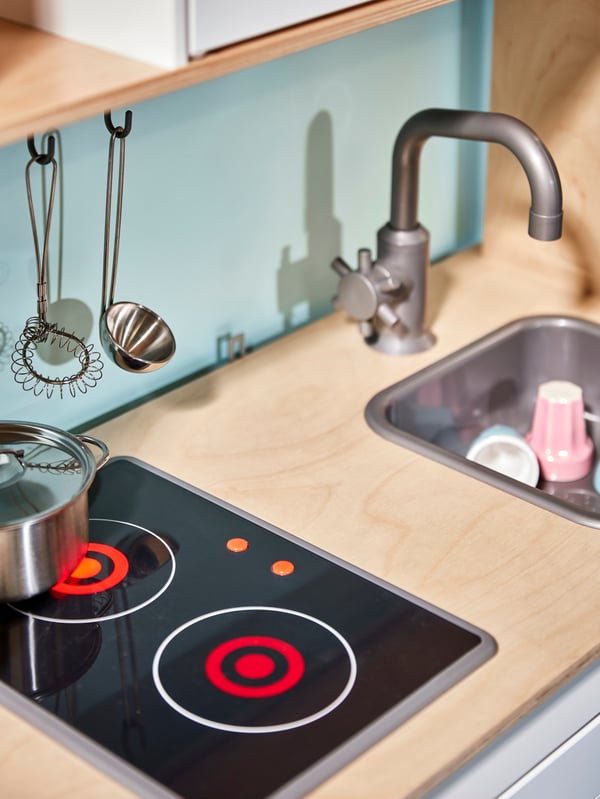 Gambar dekat dapur kanak-kanak DUKTIG yang menunjukkan sink dengan koleh di dalamnya serta hob dengan periuk bertangkai dan senduk.