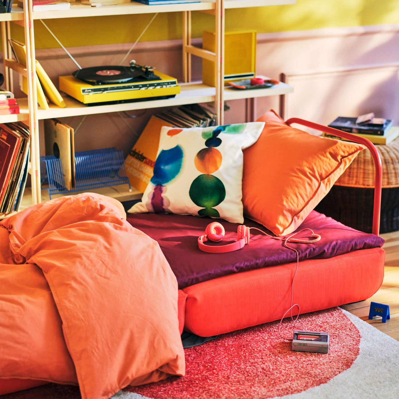 Bela IKEA PS 2026 fotelja na razvlačenje nameštena kao krevet s narandžastim jorganom, iza koje se nalaze drvene police na kojima su knjige i gramofon.