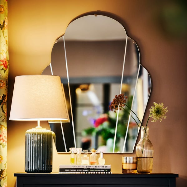 A LOMMARYD mirror is attached to the wall above a cabinet that has a lamp, books, perfume bottles and flowers on top of it.