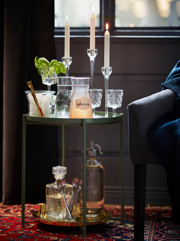 Una mesita de bar con dos jarrones con líquido adentro, al costado izquierdo de la mesa una hielera de coctel. También hay velas y una planta sobre la mesa.