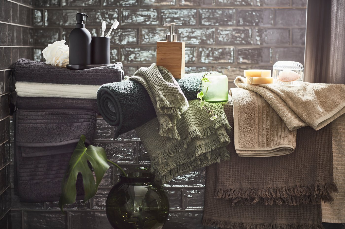 Des serviettes blanches, beige clair, brun/gris clair, gris foncé et vertes ainsi que divers accessoires dans une salle de bain aux carreaux gris.