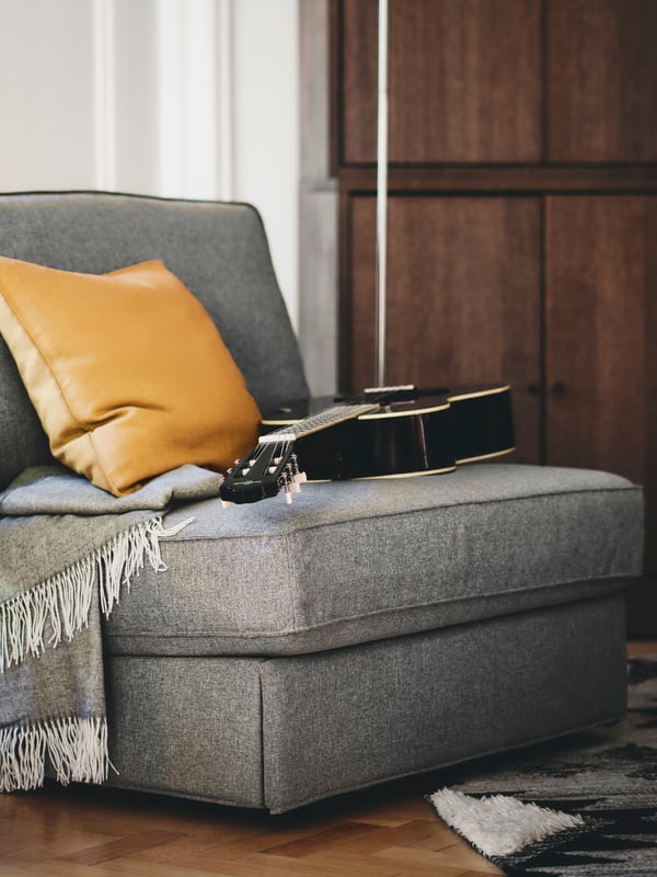 Fauteuil convertible KIVIK gris dans une pièce aux murs en bois foncé. Sur le fauteuil se trouvent une guitare, un coussin et une couverture.