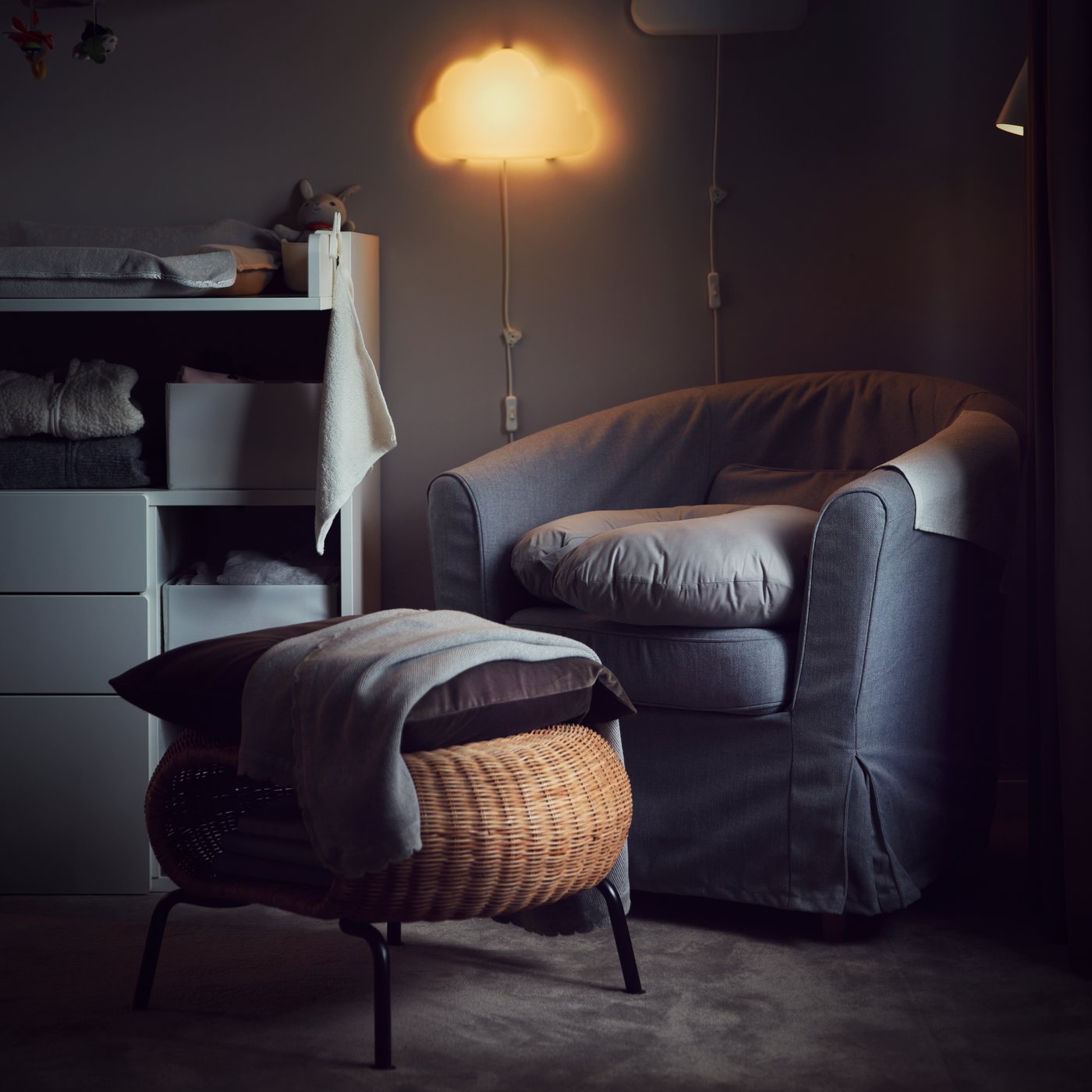 Ein Raum, der von einem Wandnachtlicht erhellt wird. Auf einem GAMLEHULT Hocker aus Rattan mit Aufbewahrung liegen Kissen und eine Decke, dahinter steht ein Sessel.