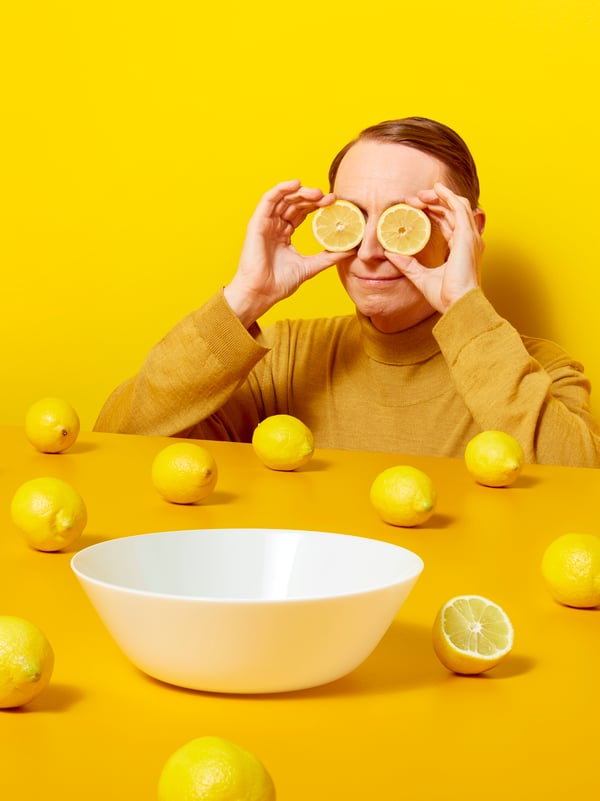 A man wearing yellow holds lemon slices over his eyes next to a yellow table with whole lemons and a white bowl on it.