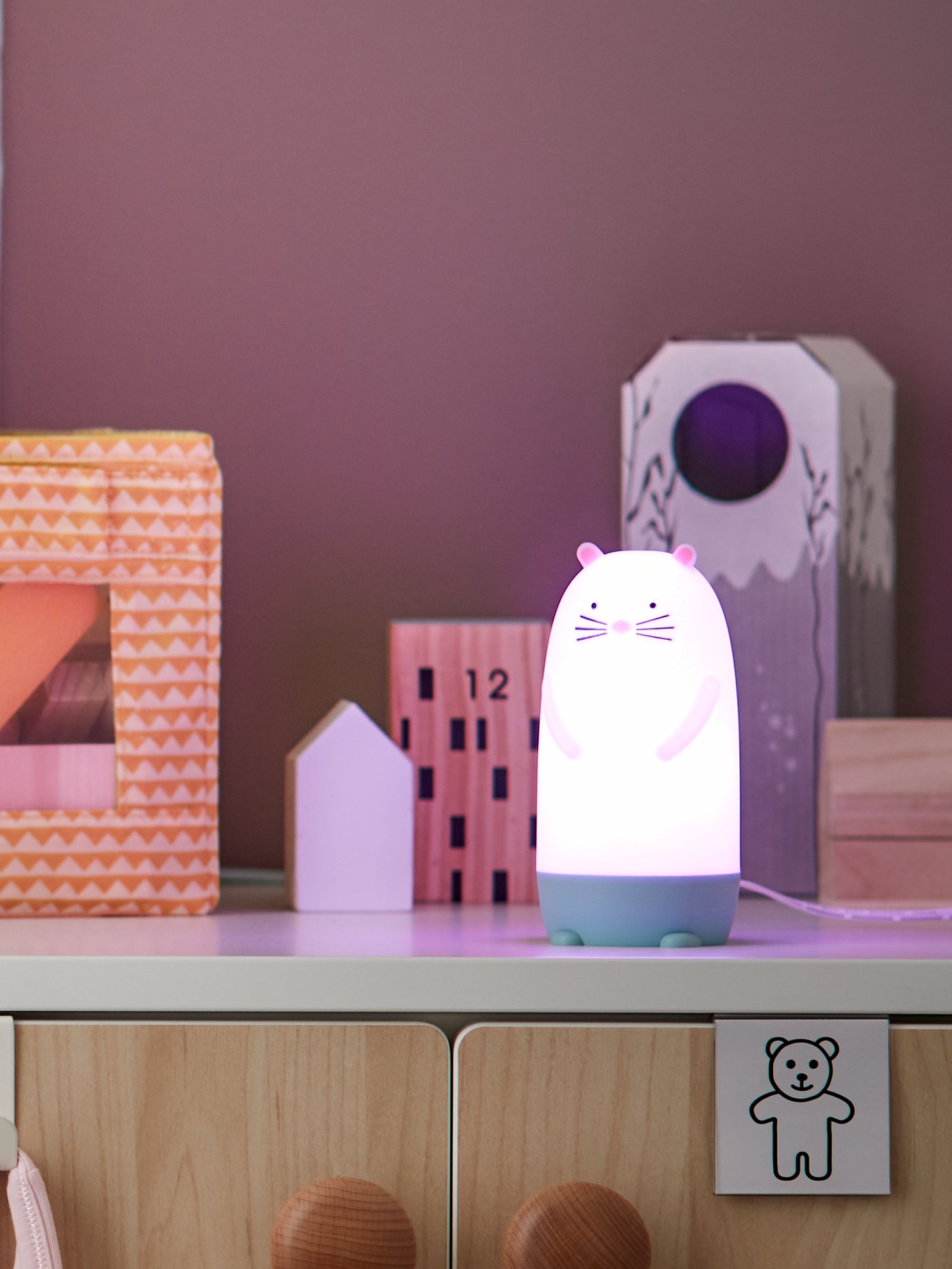 A man reads a children’s book to a young child sitting on a sofa beside a PEKHULT soft toy with LED night light.