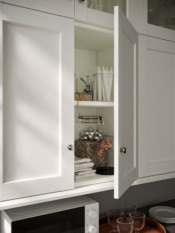 An open kitchen cabinet with ENKÖPING white doors, on the shelves are candles, towels and a KORKEN jar with lid.