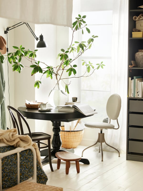 Une table à manger INGATORP noire près d’une fenêtre avec des livres, une tasse et un vase sur le dessus, et une chaise pivotante BJÖRKBERGET placée devant.