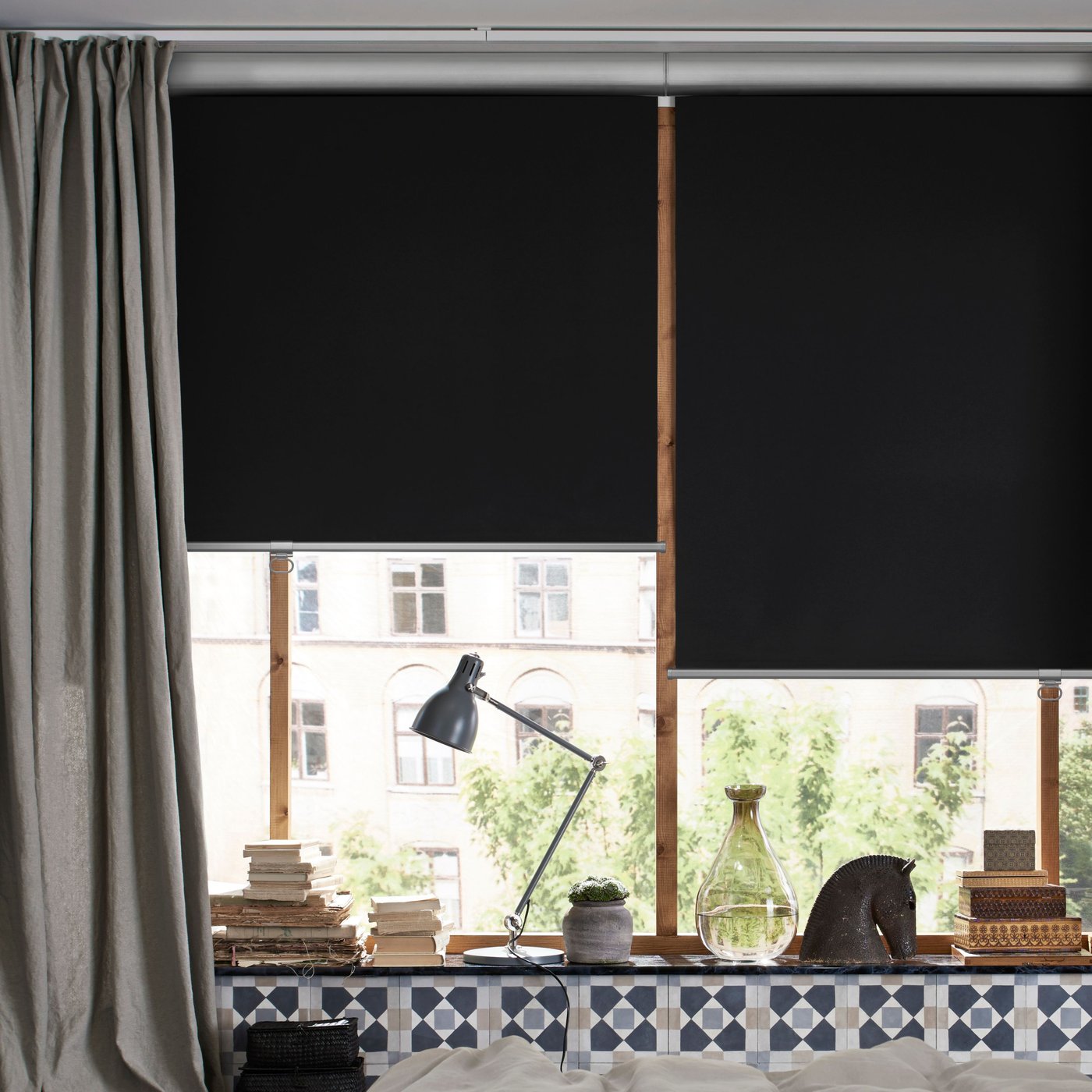 Deux stores occultants TRETUR gris foncé sur une grande fenêtre au fond d’une chambre beige, avec une lampe de bureau sur l’appui de fenêtre.
