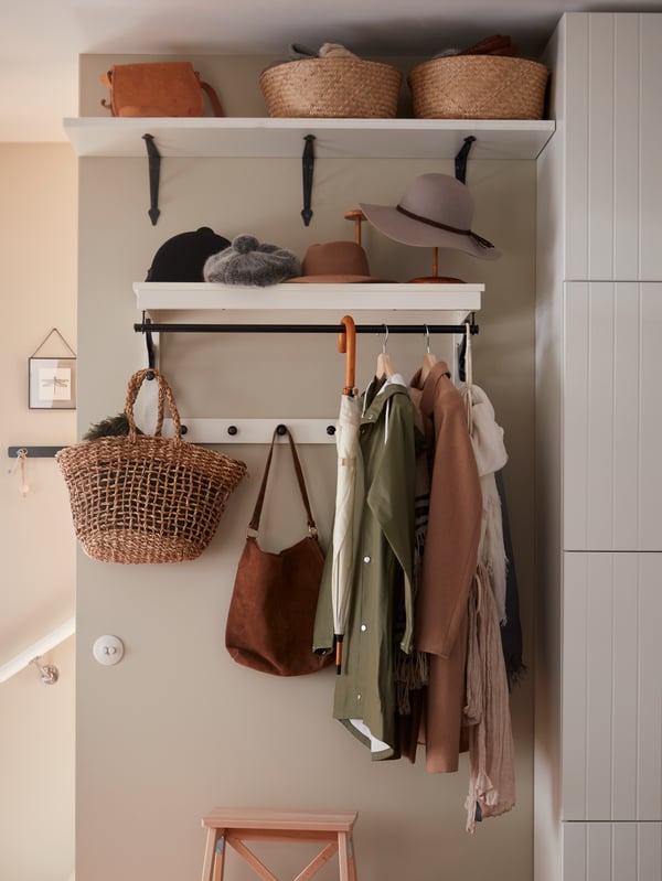 An einer beigefarbenen Wand hängt eine klassische Wandgarderobe, an der Jacken, eine Tasche, ein Schirm, ein Korb und Hüte zu sehen sind