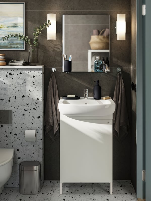 A NYSJÖN cabinet with one door stands under a white sink with light grey-blue accessories on top and a mirror on the wall.