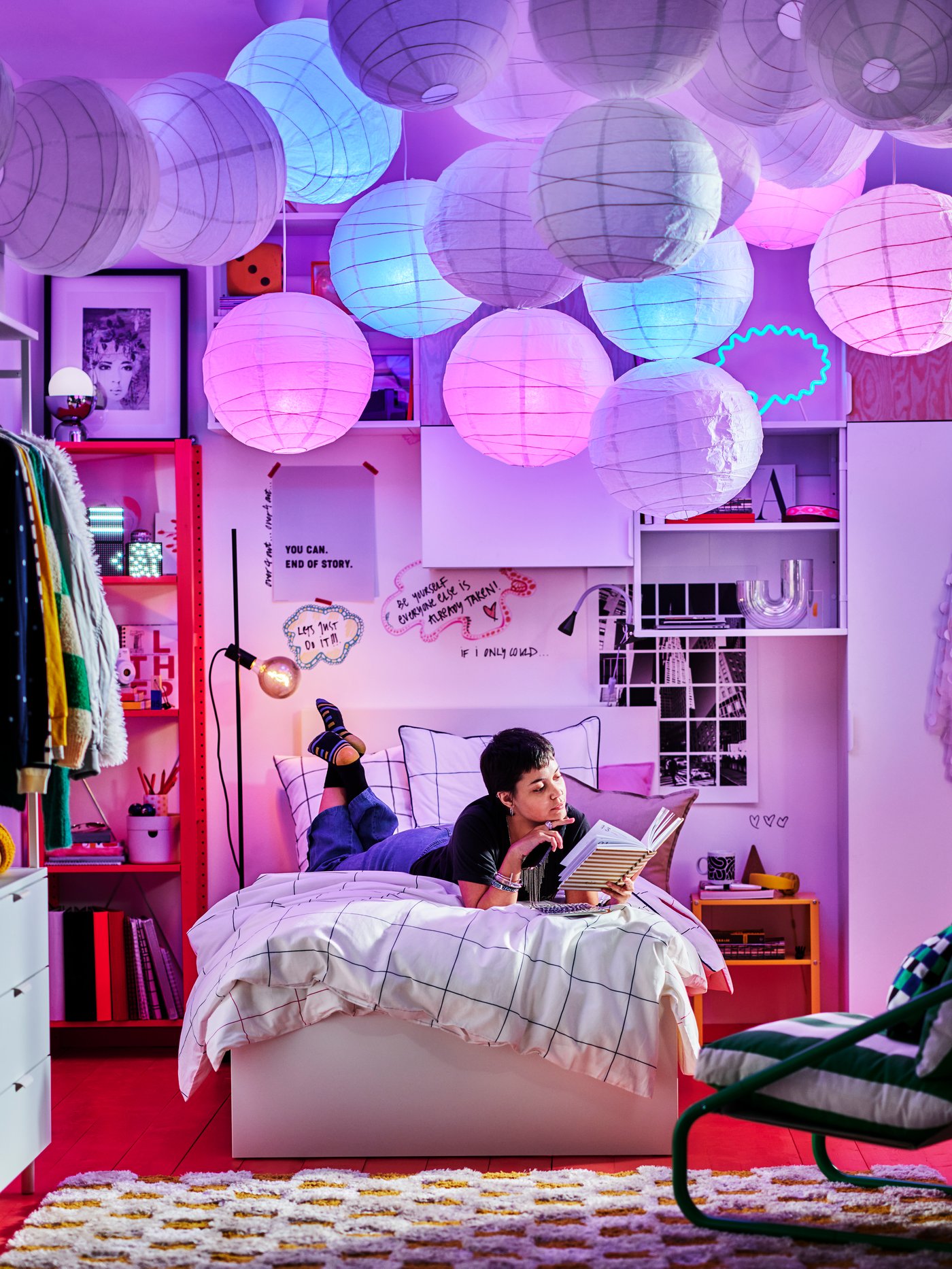 A girl is reading a book while laying on her bed. On top of her, many colorful decorations are attached to the room's ceiling