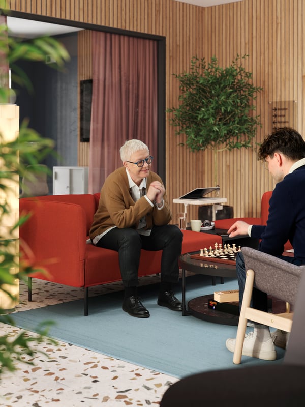 Crvena LILLEHEM modularna sofa na kojoj sedi osoba koja čeka da protivnik u šahu povuče potez nalazi se u modernom lobiju s drvenim panelima.
