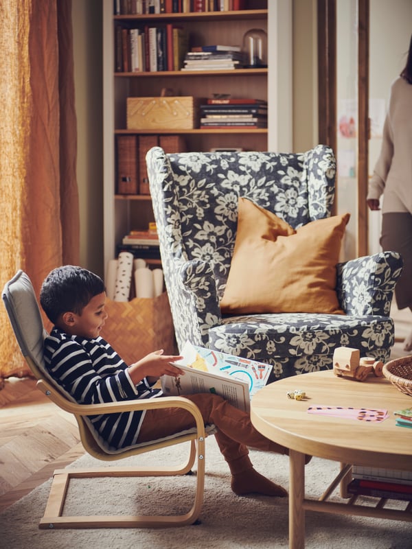 Un niño sentado en un sillón infantil POÄNG junto a un sillón orejero STRANDMON con una funda extraíble de estampado llamativo en un salón.