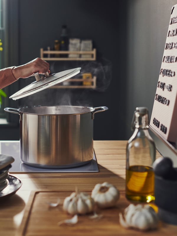 IHコンロの上のステンレススチール製IKEA 365+ スープ鍋のガラス製ふたを開けている人。そのそばには、竹製のAPTITLIG/アプティートリグ まな板。