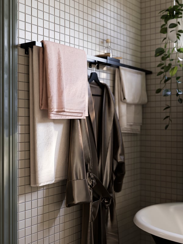 A tiled bathroom wall with SKOGSVIKEN towel rails in black holding VINARN bath towels in pink and light grey/beige.