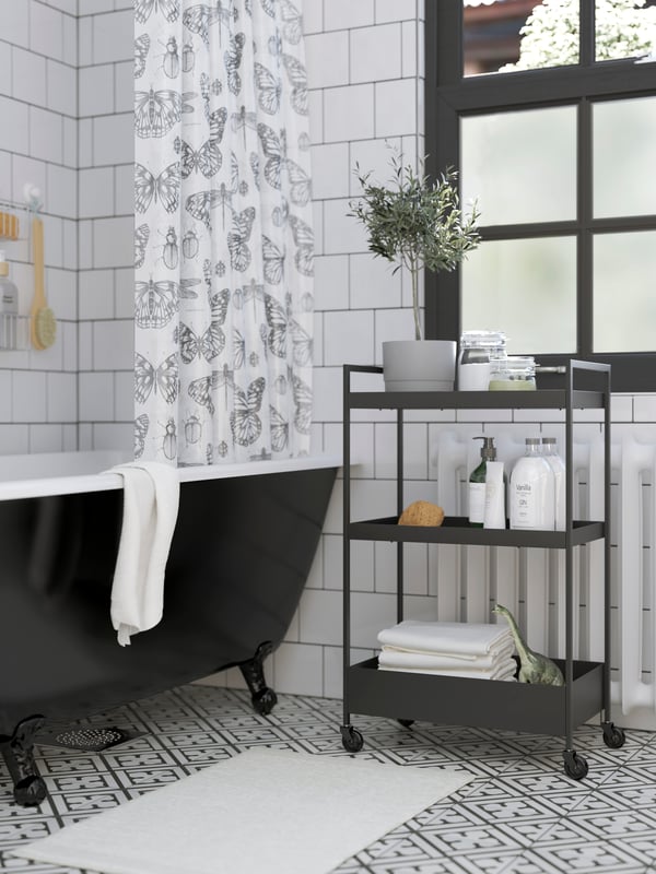 Carrello nero con asciugamani e prodotti per la doccia, vasca da bagno nera, tappeto per bagno bianco e tenda doccia con disegni di farfalle.