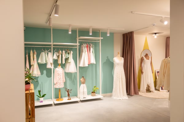 Een stijlvolle winkelruimte met een groene wand, lichte kleding op hangrekken, paspoppen in jurken en zachte verlichting die een rustige sfeer creëert.