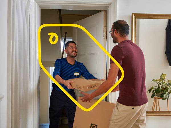 Hymyilevä kotiinkuljetuspalvelun mies ojentaa paketteja miehelle, joka seisoo asuntonsa eteisessä.