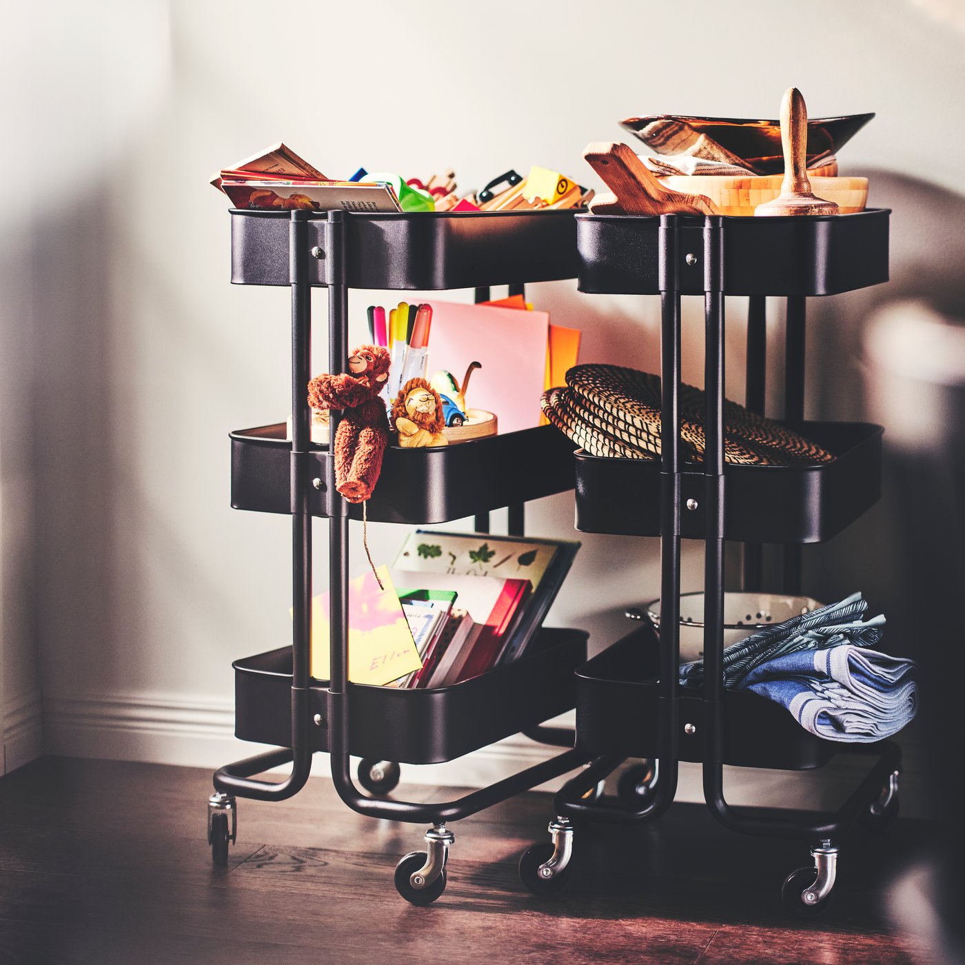 Dos carros negros RÅSKOG que transportan juguetes, libros, plumas, dibujos, paños y algunos manteles individuales LÄTTAD hechos de hierba marina.
