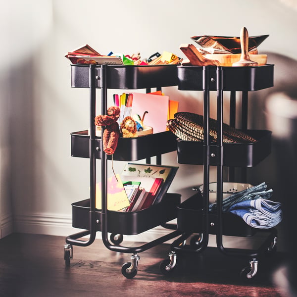 Two black RÅSKOG carts carrying toys, books, pens, drawings, cloths and a few seagrass LÄTTAD placemats.