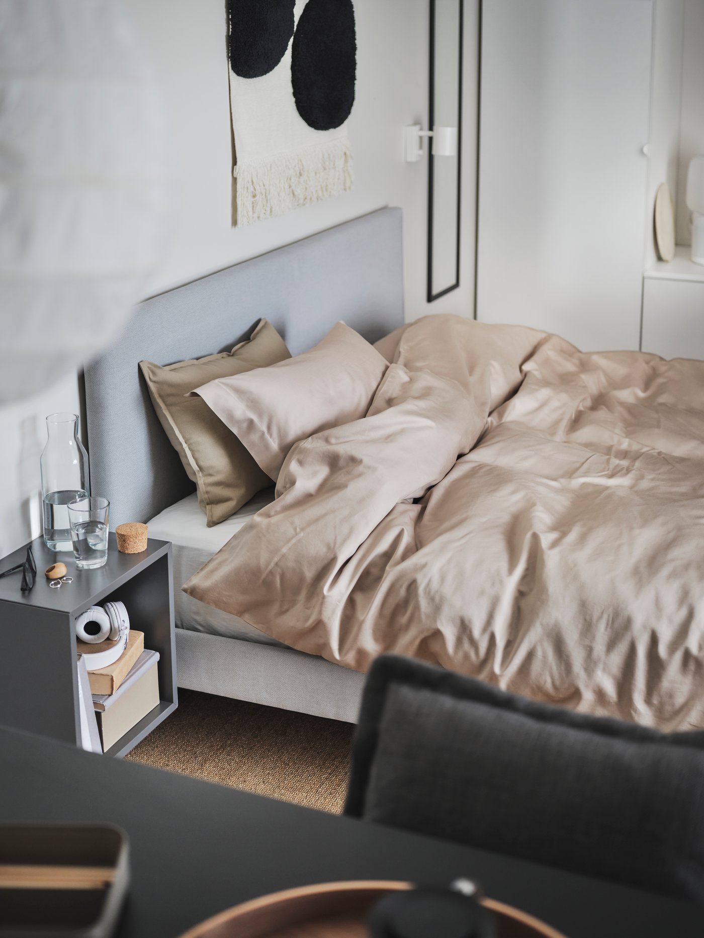 Modernes Schlafzimmer mit einem weißen Bettgestell und einem hellgrauen Kopfteil, bezogen mit einem glänzenden, beige-goldenen Bettwäsche-Set. Daneben steht ein kleiner Nachttisch mit Dekoration, im Vordergrund ein schwarzer Tisch mit Geschirr. Die Wände sind hell und minimalistisch gestaltet.