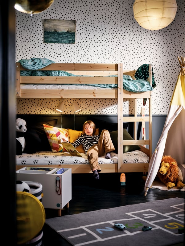 Une chambre noir et blanc où un petit garçon a le dos appuyé au mur sur le lit du bas du lit superposé MYDAL, entourés de plusieurs jouets.