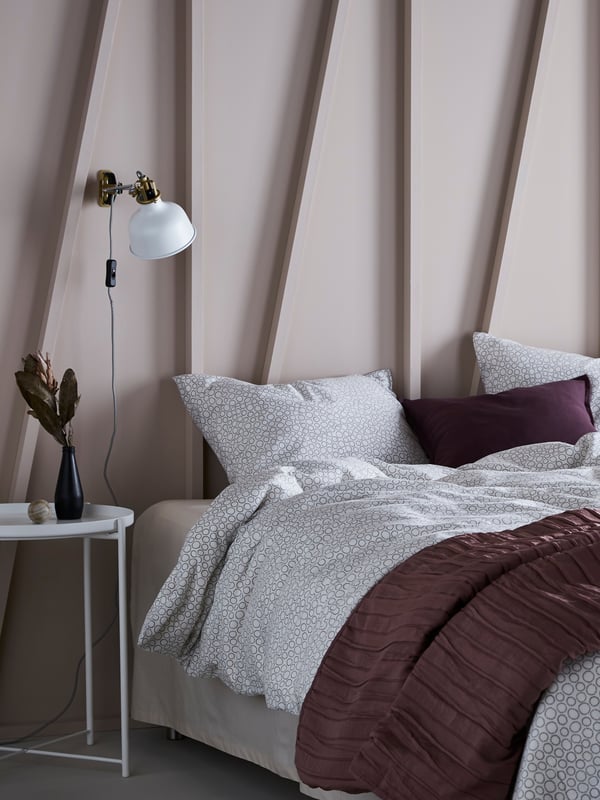 A neatly made bed with a light purple bed behind it and a RANARP white lamp attached on the wall next to the bedside