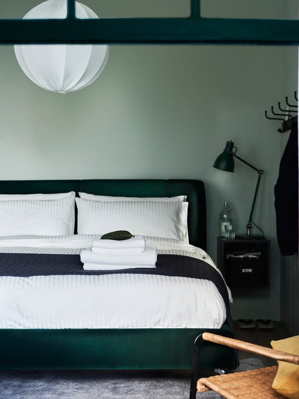 A bedroom with a TUFJORD dark green upholstered bed frame and white bed linen with a dark throw folded across the end.