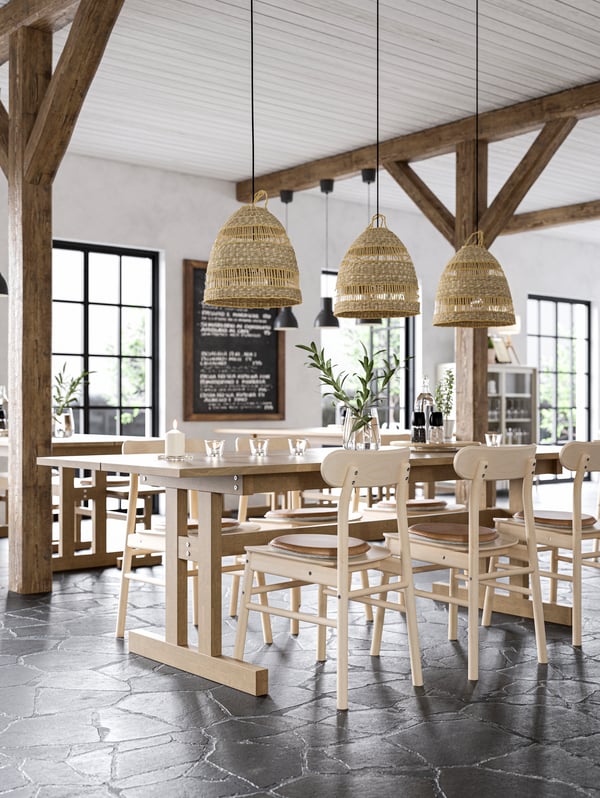 Un restaurante con diseño estilo cabaña con amplias mesas y sillas de madera. Sobre las mesas cuelgan lámparas rústicas y a los costados se ven grandes vigas de madera.