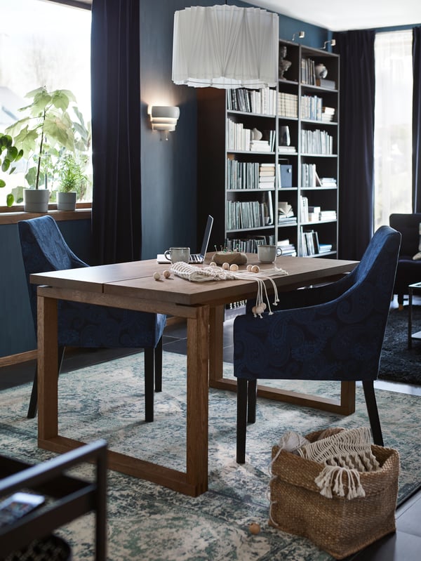 A wooden table with two blue dining chairs around it, a laptop and craft materials on top, and a white pendant lamp above.