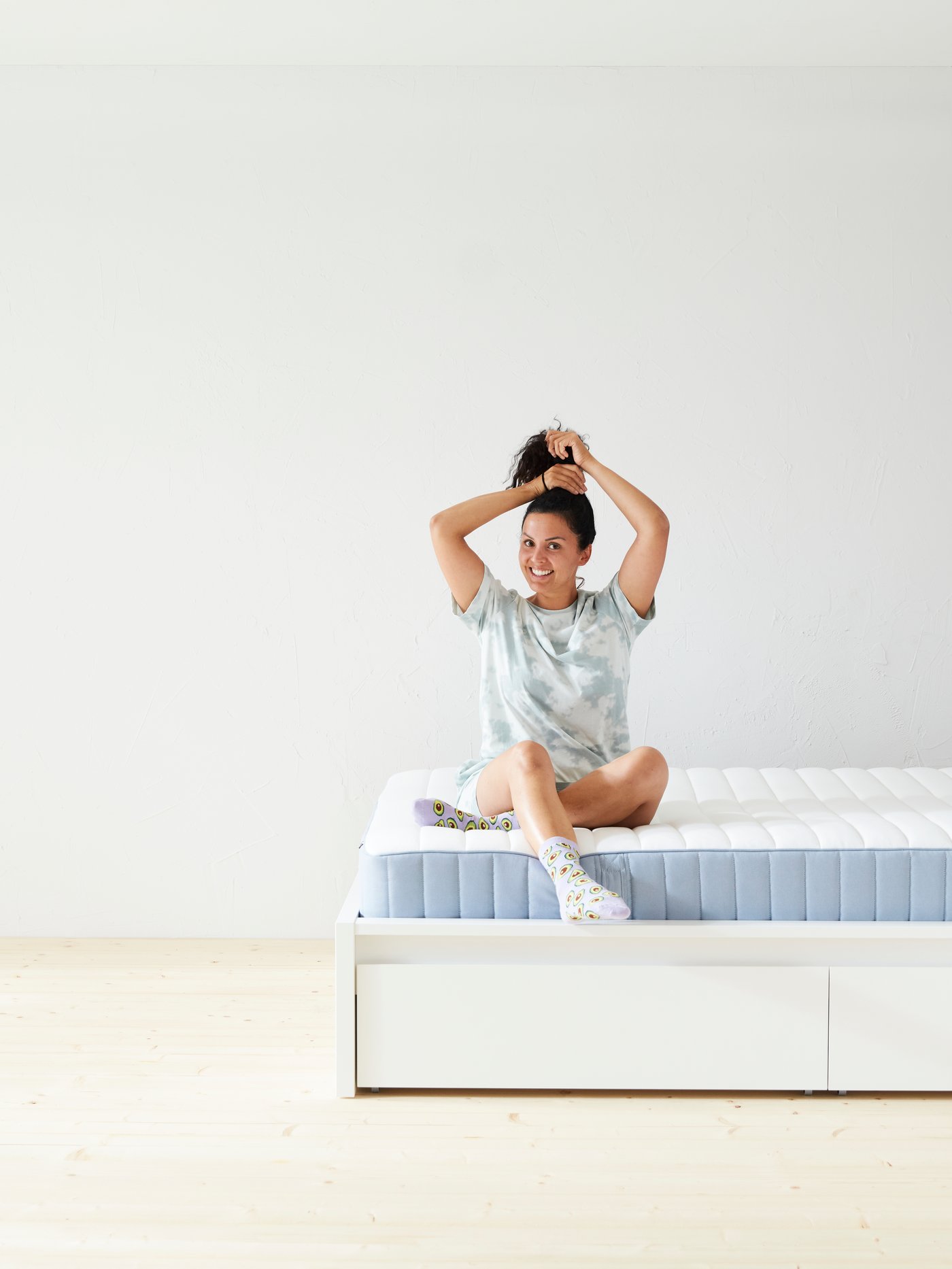 A woman sits on her bed with a smile on her face and ties her hair up in a high ponytail