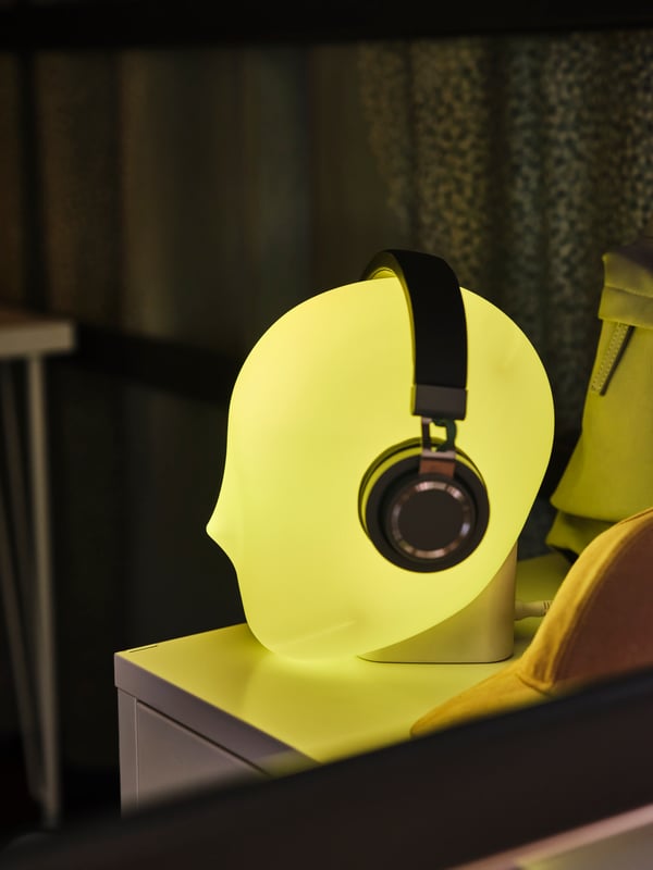 An ISKÄRNA LED table lamp in a shape of a head shining in yellow colour with headphones on in a dark room.