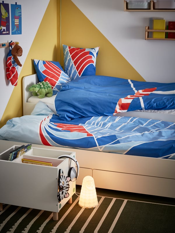 Una cama nido SLÄKT con cajones de almacenamiento abajo, cubierta por sábanas SPORTSLIG en colores azul, celeste y rojo. Frente a la cama hay un mueble para almacenamiento SMUSSLA y una lámpara de noche SOLBO. 