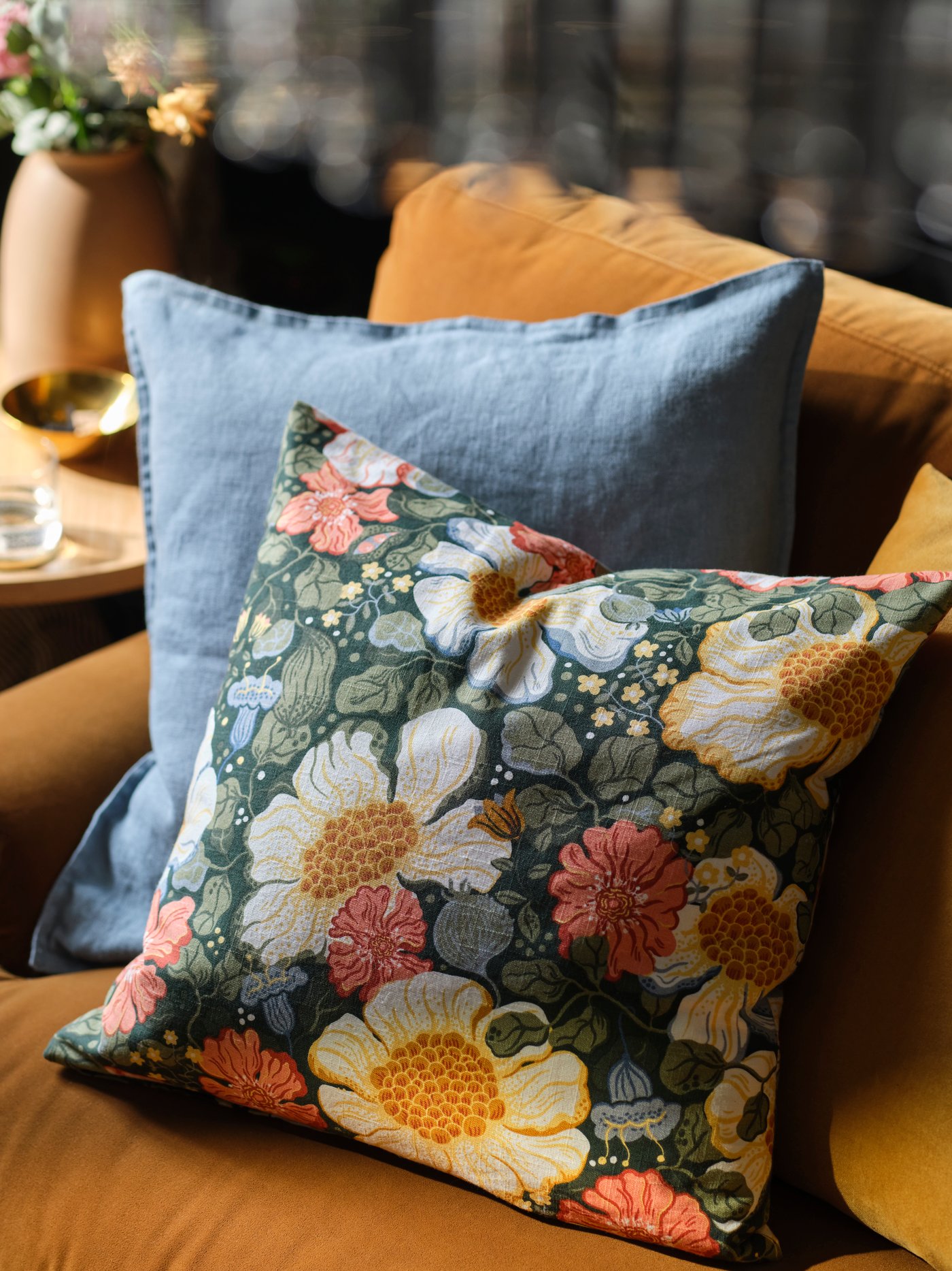 A couple of cushions lean on a yellow-brown sofa, one is dressed in an ARENBOK cushion cover with a floral pattern.
