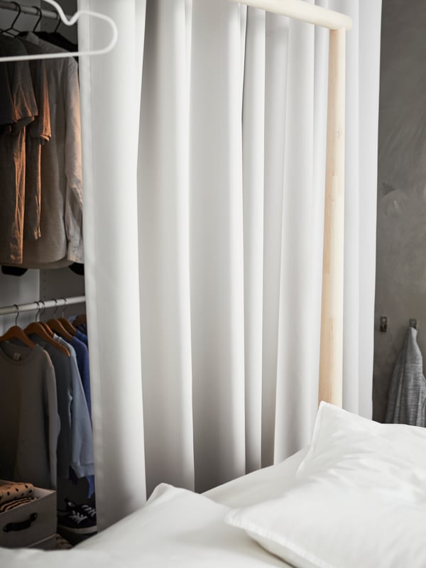 Light gray MAJGULL room darkening curtains, used as a divider between a closet full of clothes and a birch GJÖRA bed frame.