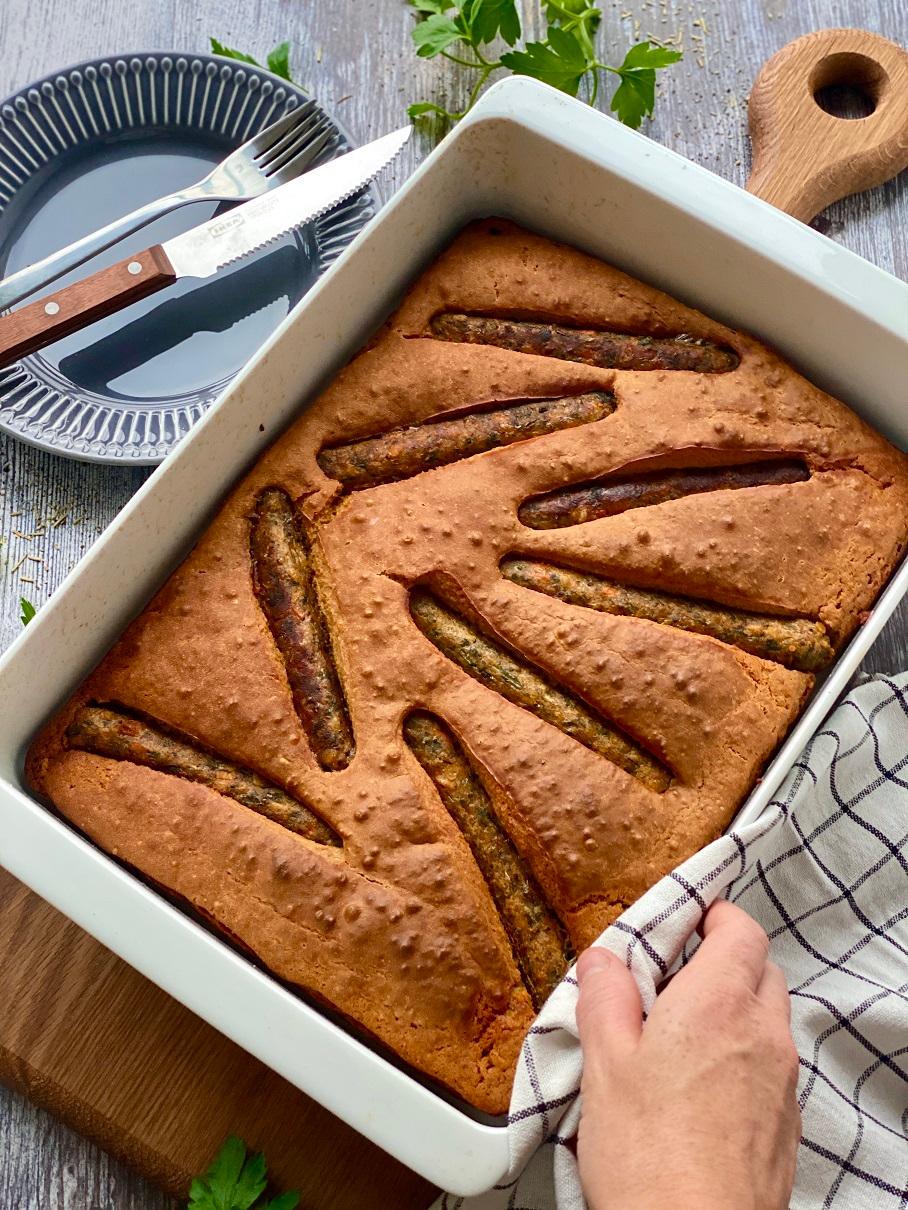 Gâteau fraîchement sorti du four, avec de fines entailles sur le dessus, présenté sur un plat blanc posé sur une planche en bois, accompagné de couverts et d'une nappe à carreaux.