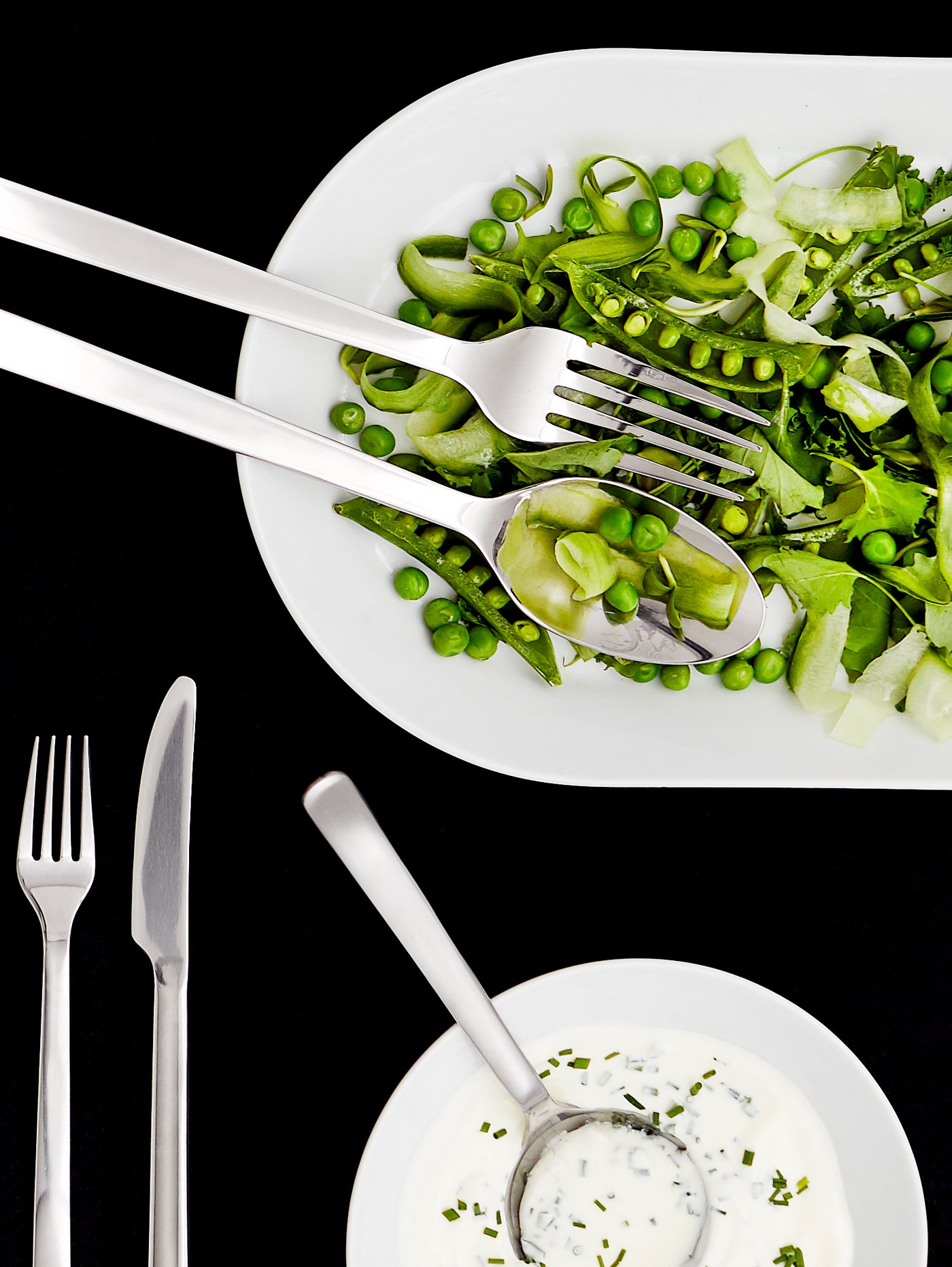 Platos y fuentes para servir blancas con cubiertos metálicos y platos con ensaladas verdes.