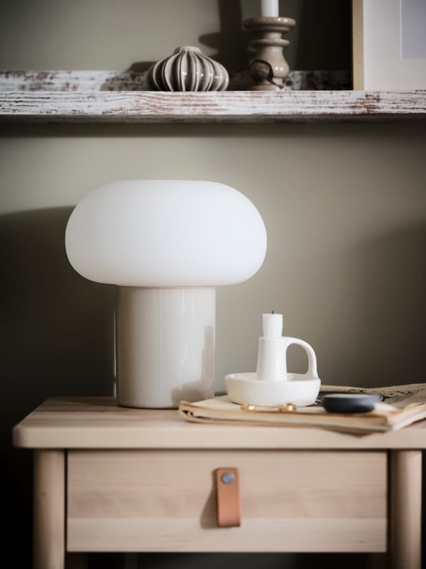 An opal-and-white DEJSA table lamp stands next to a white candlestick on a BJÖRKSNÄS bedside table.

