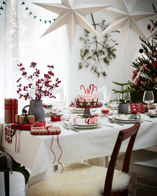 Table festive dressée avec une nappe blanche et des éléments de décoration VINTERFINT rouges. Un abat-jour blanc STRÅLA est suspendu au plafond. Au premier plan se trouve une chaise PINNTORP marron avec une fourrure pour une assise moelleuse.
