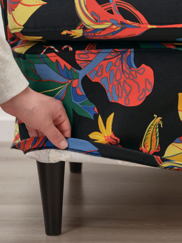 A person’s hand holds the edge of a Stigsbo multicolour/black slipcover for wing chair covering a STRANDMON wing chair.