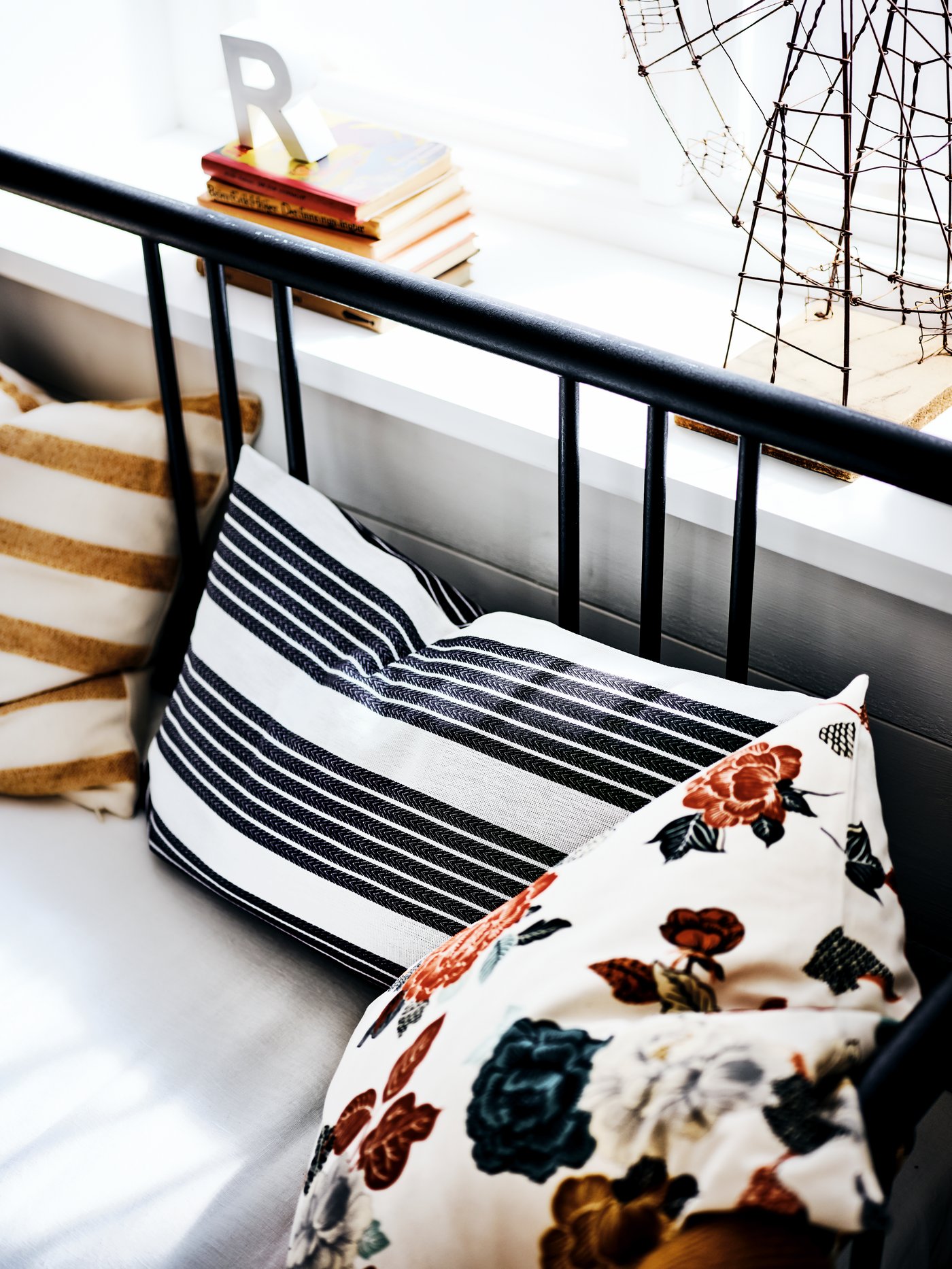 La structure métallique noire du lit banquette devant une fenêtre avec des coussins sur le lit, à rayures et à motifs de fleurs.