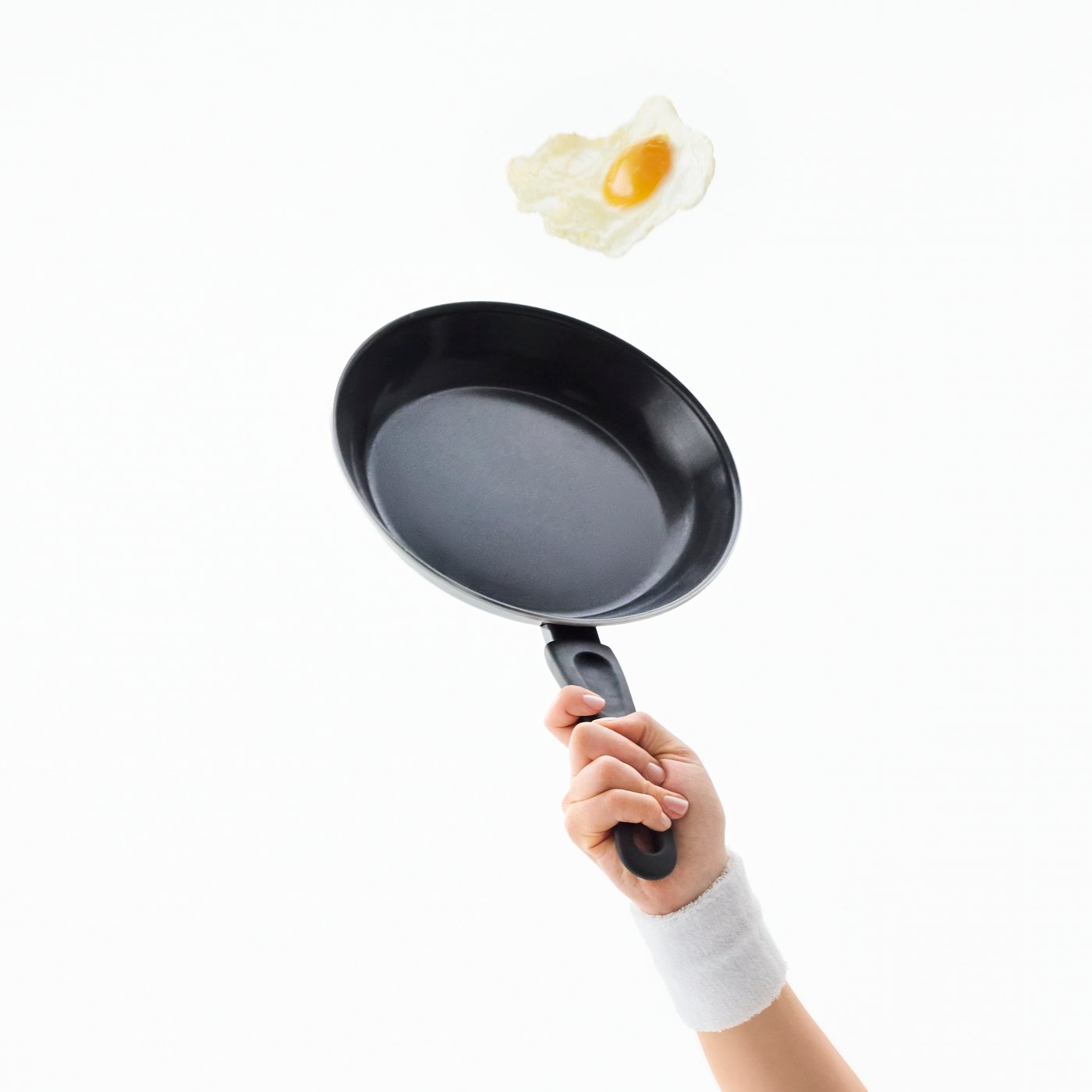 An arm is holding a TAGGHAJ frying pan as if it were a tennis racquet, catching a fried egg in the air as if it were a ball.