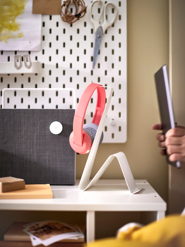 A tablet on a black MÖJLIGHET headset and tablet stand on a white desk with headphones hanging from it.