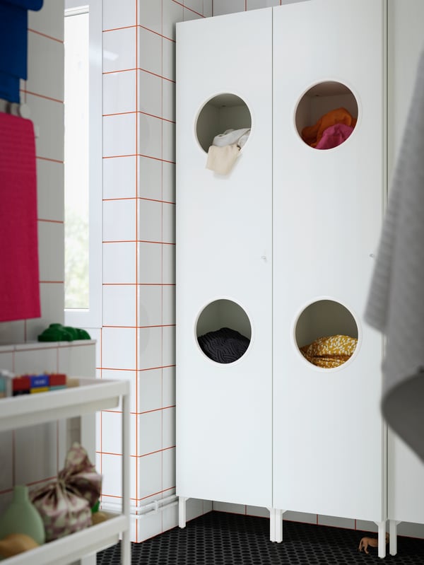 A NYSJÖN laundry cabinet with circular openings for sorting clothes, featuring towels and clothing stored inside.