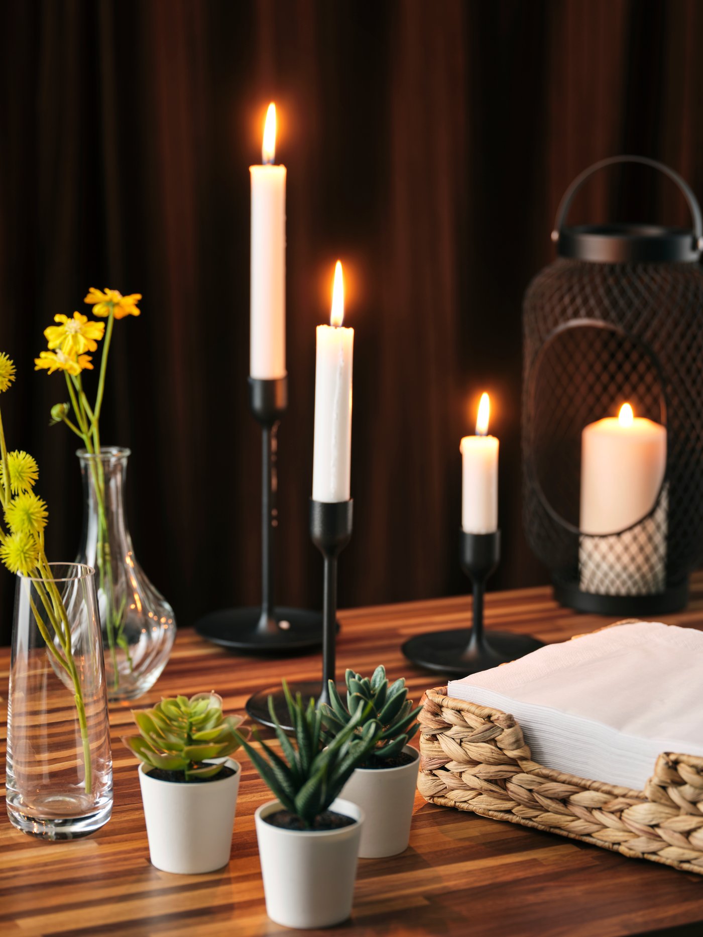 A black FULLTALIG candlestick set of three holding tall white candles, accented by small potted succulents and flowers.