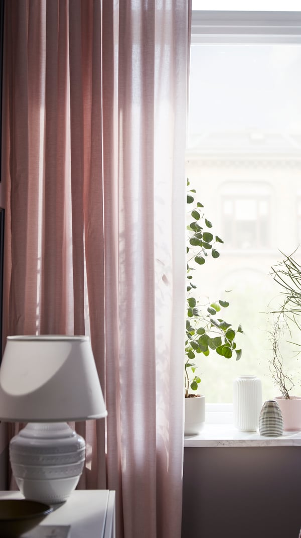 Finestra di un soggiorno con tende HANNALILL rosa scuro che lasciano filtrare la luce, alcune piante e portavasi sul davanzale - IKEA