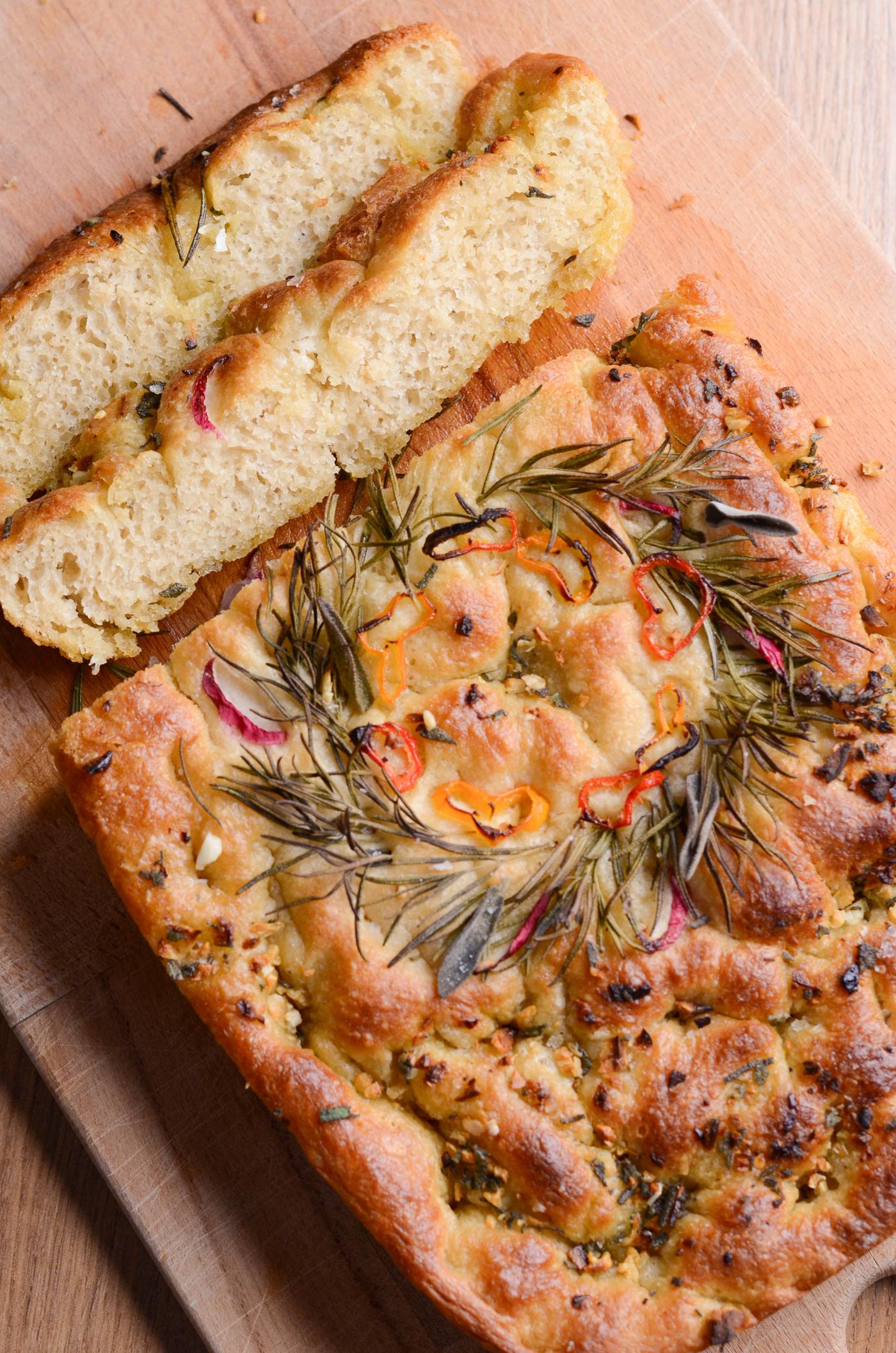 Focaccia fraîchement cuite dans un plat rectangulaire, garnie d'une couronne de romarin et de tomates. Un plat contenant de l'huile d'olive et du beurre est posé à proximité sur une table en bois.